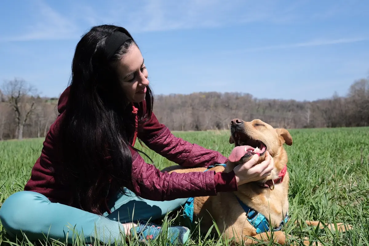 Abby sitting in a grassy field, smiling as she scratches her dog Anderson behind the ears.